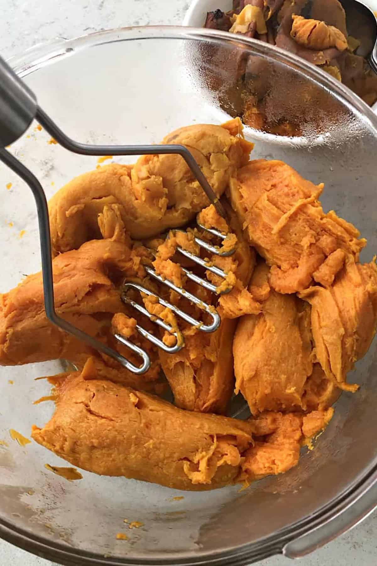 A metal potato masher presses cooked sweet potatoes in a clear glass bowl, preparing them to be mashed.