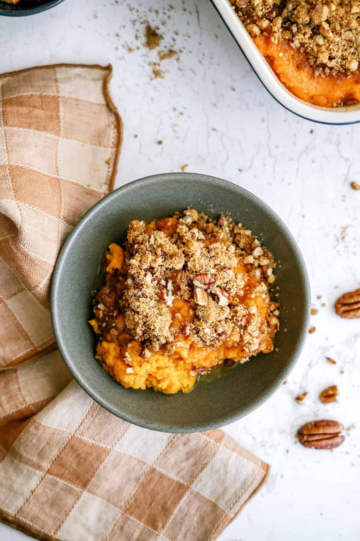 A white baking dish contains a sweet potato casserole topped with a pecan and crumb mixture; a large portion has been scooped out.