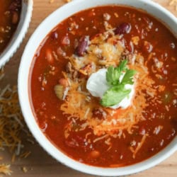 A bowl of chili topped with shredded cheese, a dollop of sour cream, and a sprig of cilantro, with scattered cheese on the wooden surface nearby.
