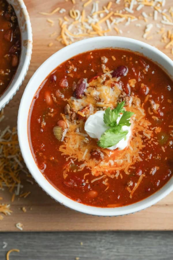 A bowl of chili topped with shredded cheese, a dollop of sour cream, and a sprig of cilantro, with scattered cheese on the wooden surface nearby.