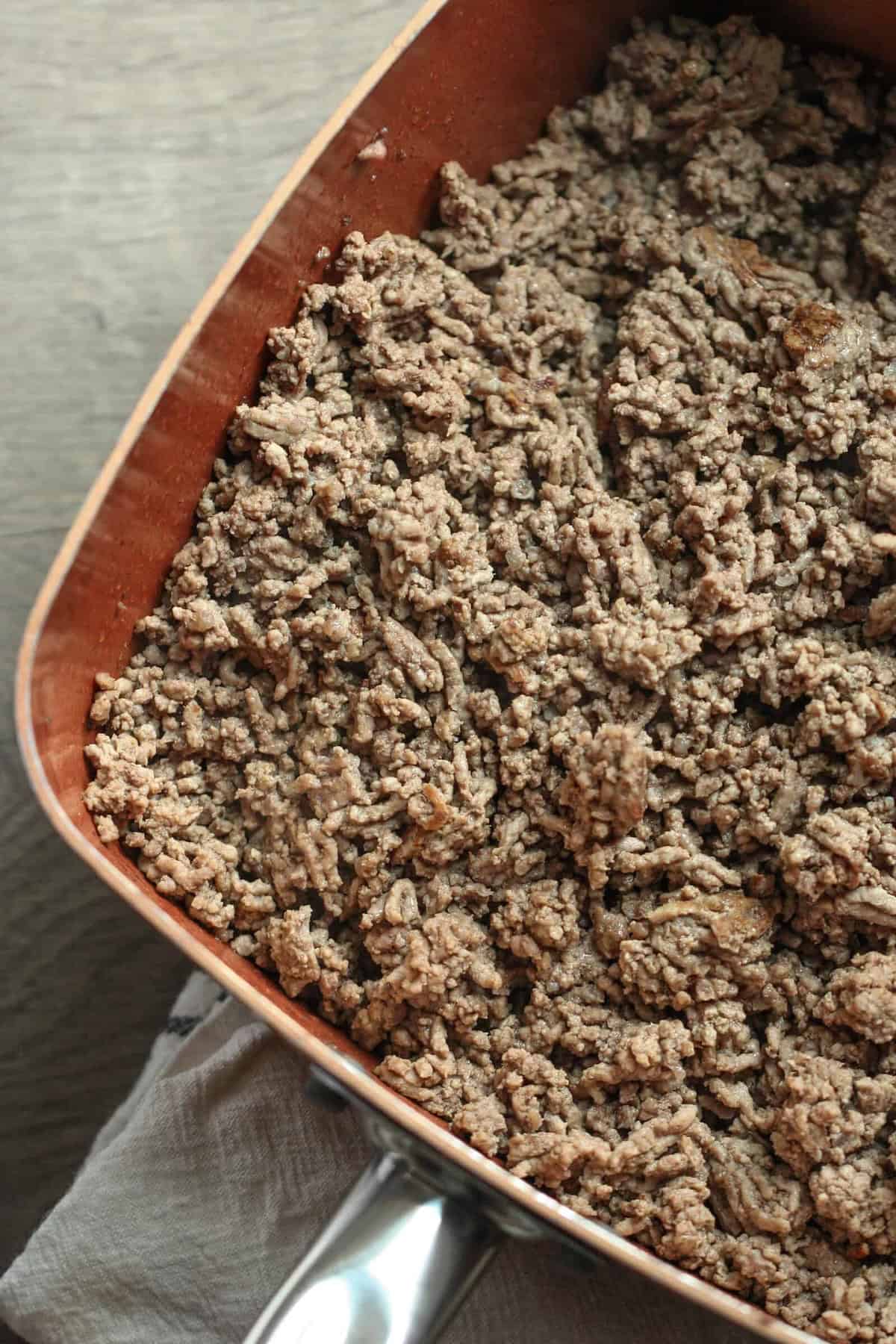 Cooked ground beef in a copper-colored skillet, resting on a light cloth, viewed from above.