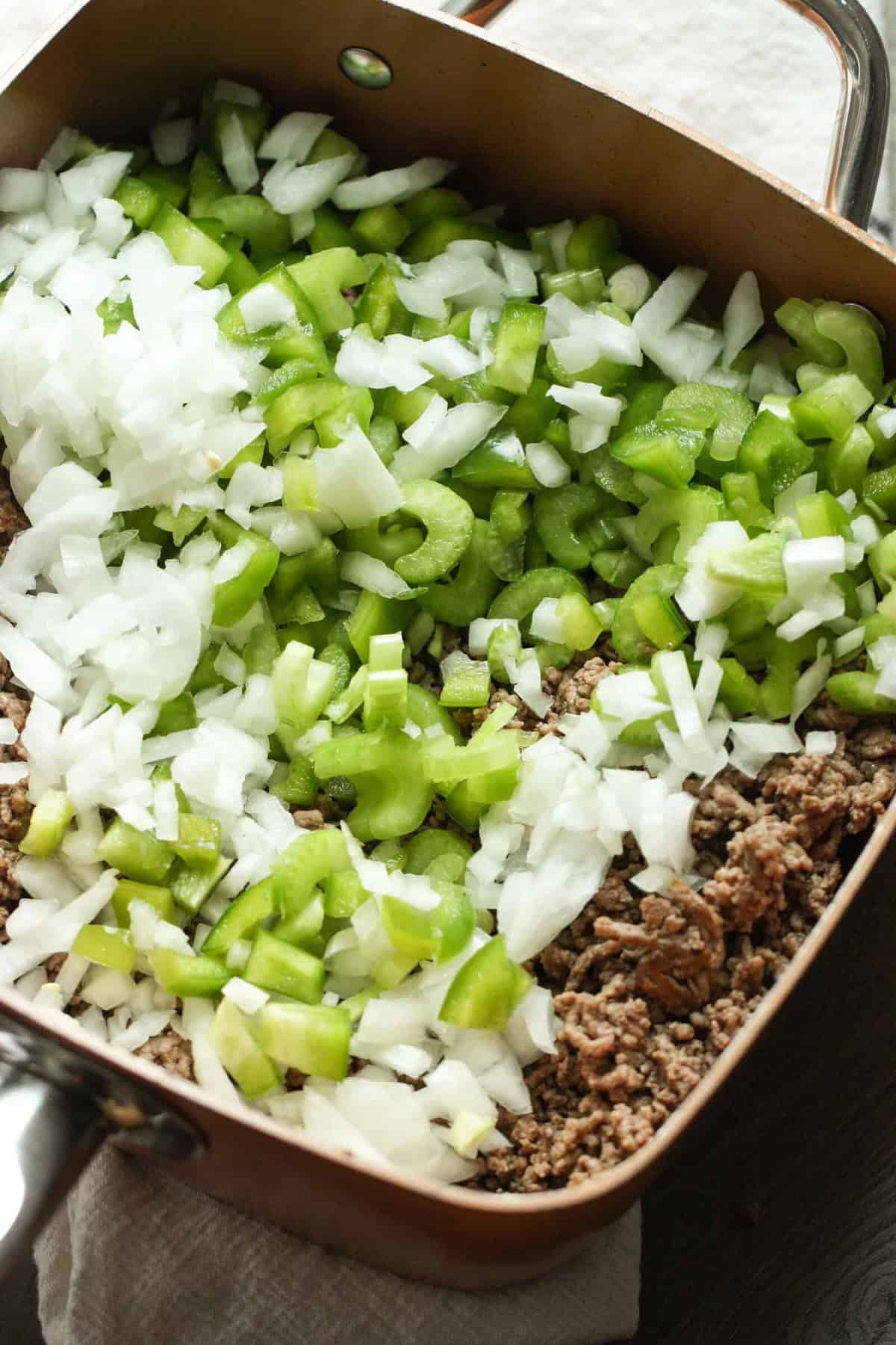 A skillet contains browned ground beef topped with a layer of diced green bell peppers and chopped onions, ready to be mixed or cooked further.