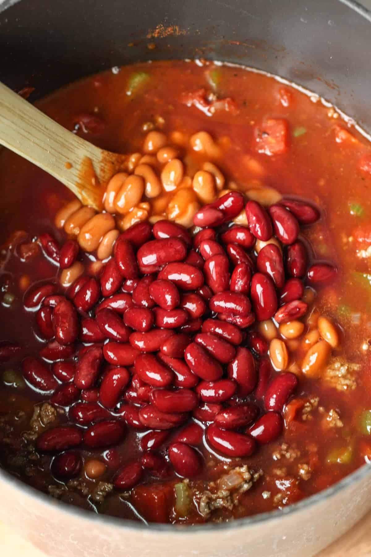A pot of chili with kidney beans, pinto beans, ground meat, tomatoes, and bell peppers being stirred with a wooden spoon.