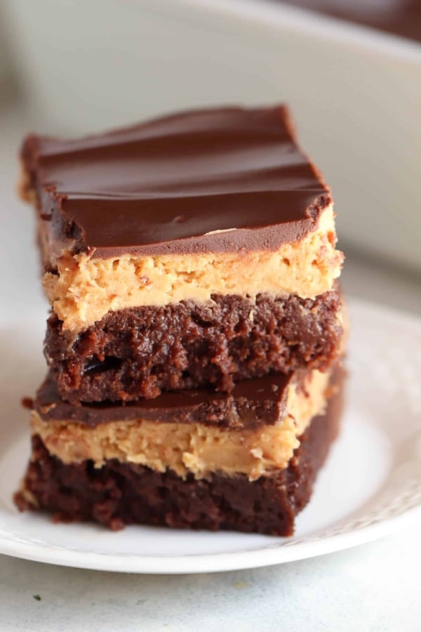 A close-up of two stacked chocolate peanut butter brownies on a white plate. The brownies have a thick, rich layer of chocolate frosting on top.