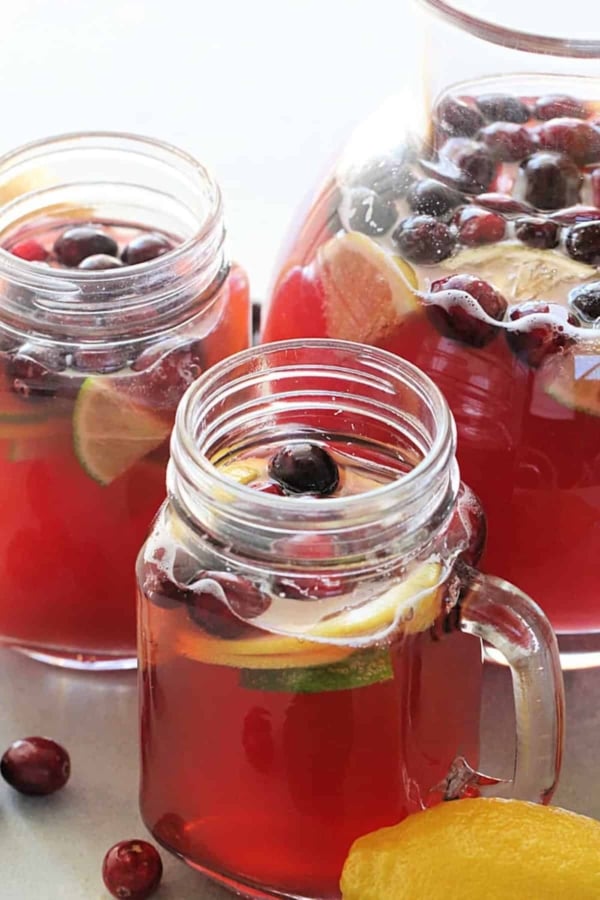 Glass pitcher and two mason jar mugs filled with cranberry beverage, garnished with lemon and lime slices, and cranberries.