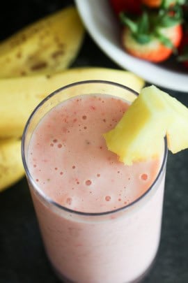 A glass filled with a pink smoothie, garnished with a pineapple slice, and bananas in the background.