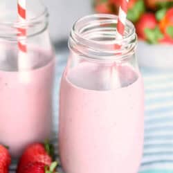 Two mason jars filled with pink strawberry smoothies and striped straws, with fresh strawberries placed nearby on a blue and white striped cloth.