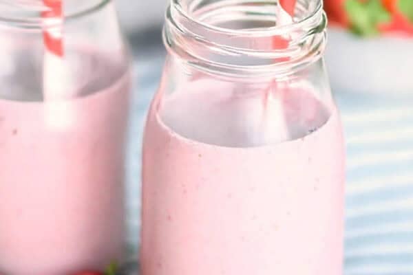 Two mason jars filled with pink strawberry smoothies and striped straws, with fresh strawberries placed nearby on a blue and white striped cloth.