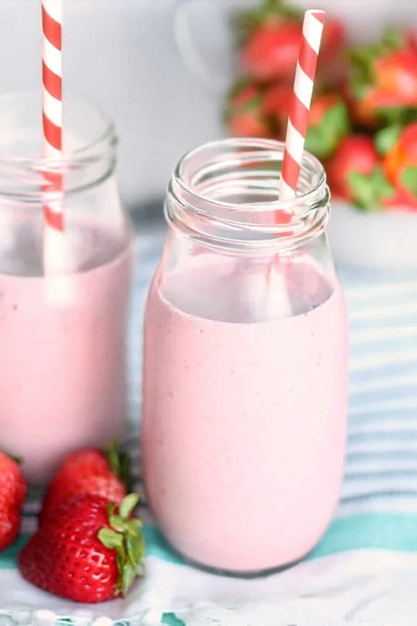 Two mason jars filled with pink strawberry smoothies and striped straws, with fresh strawberries placed nearby on a blue and white striped cloth.