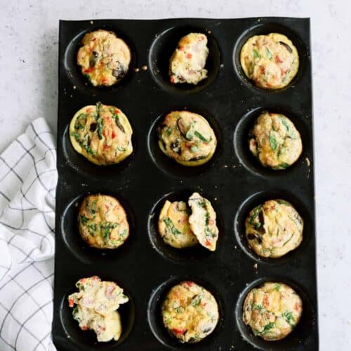 Muffin tray with twelve cooked egg muffins, some with spinach and vegetables, on a light surface next to a checkered cloth.
