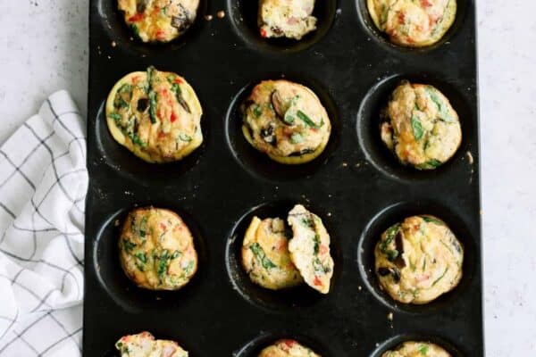 Muffin tray with twelve cooked egg muffins, some with spinach and vegetables, on a light surface next to a checkered cloth.