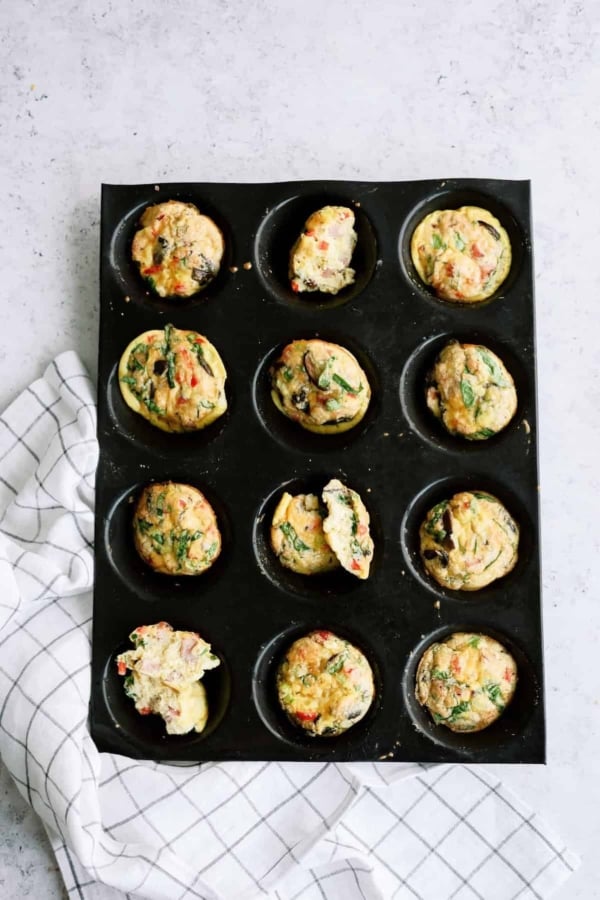 Muffin tray with twelve cooked egg muffins, some with spinach and vegetables, on a light surface next to a checkered cloth.
