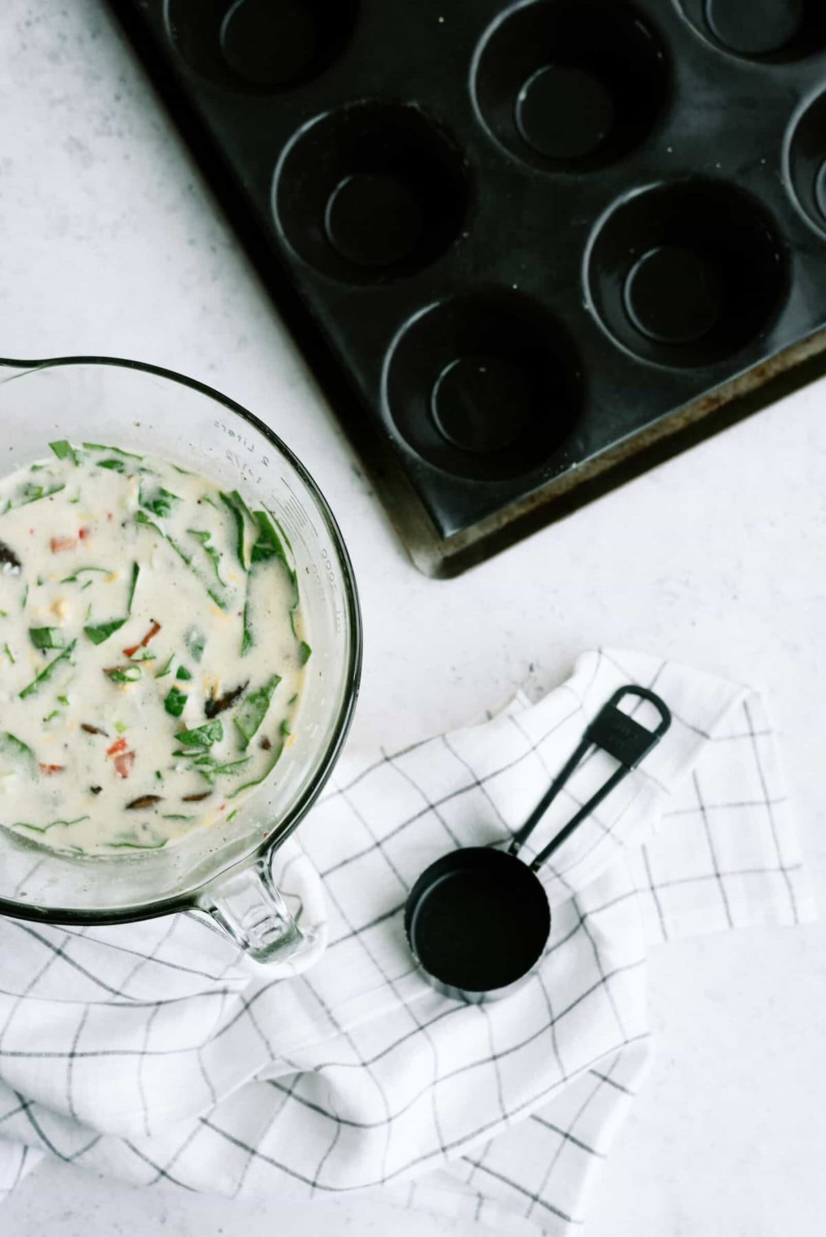 Mixing bowl with batter, a checkered cloth, measuring spoons, and a muffin pan on a white surface.
