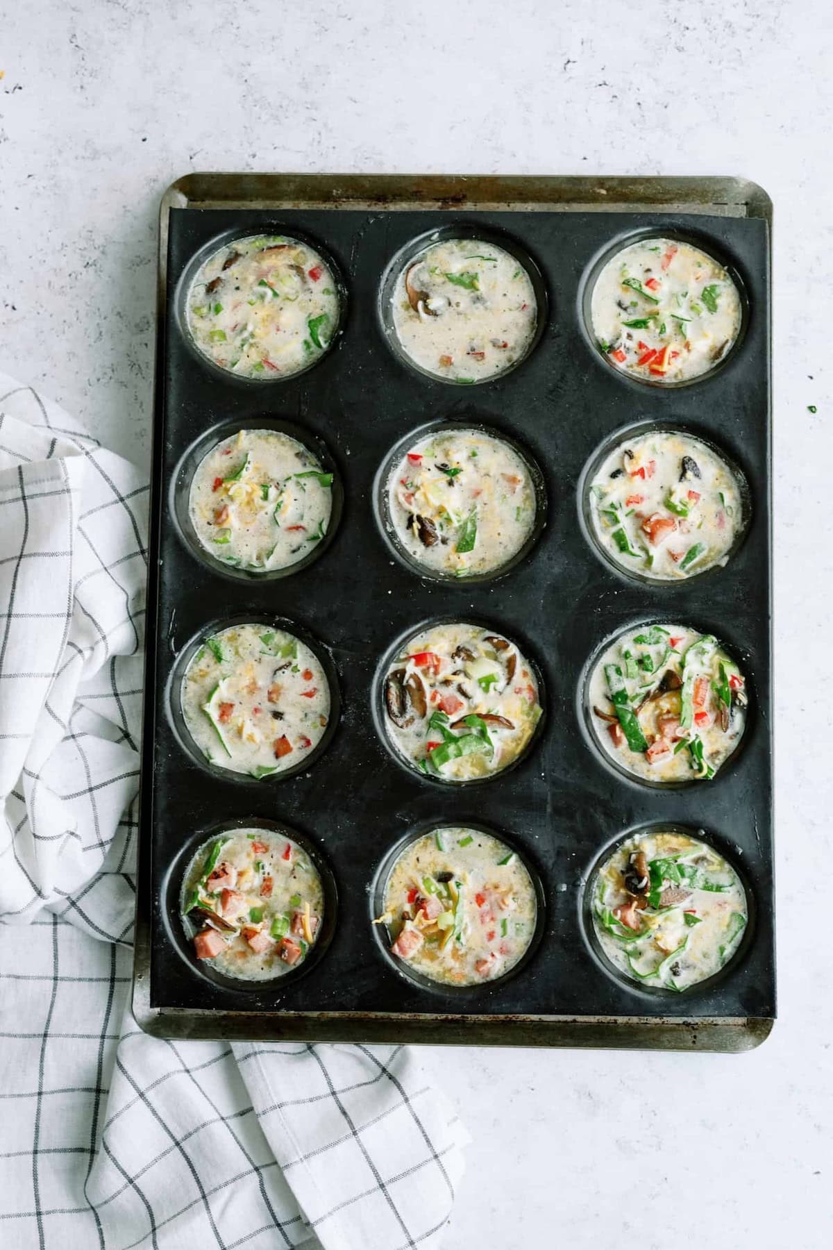Muffin tray filled with uncooked egg mixture containing vegetables and herbs, next to a white and black checkered cloth.