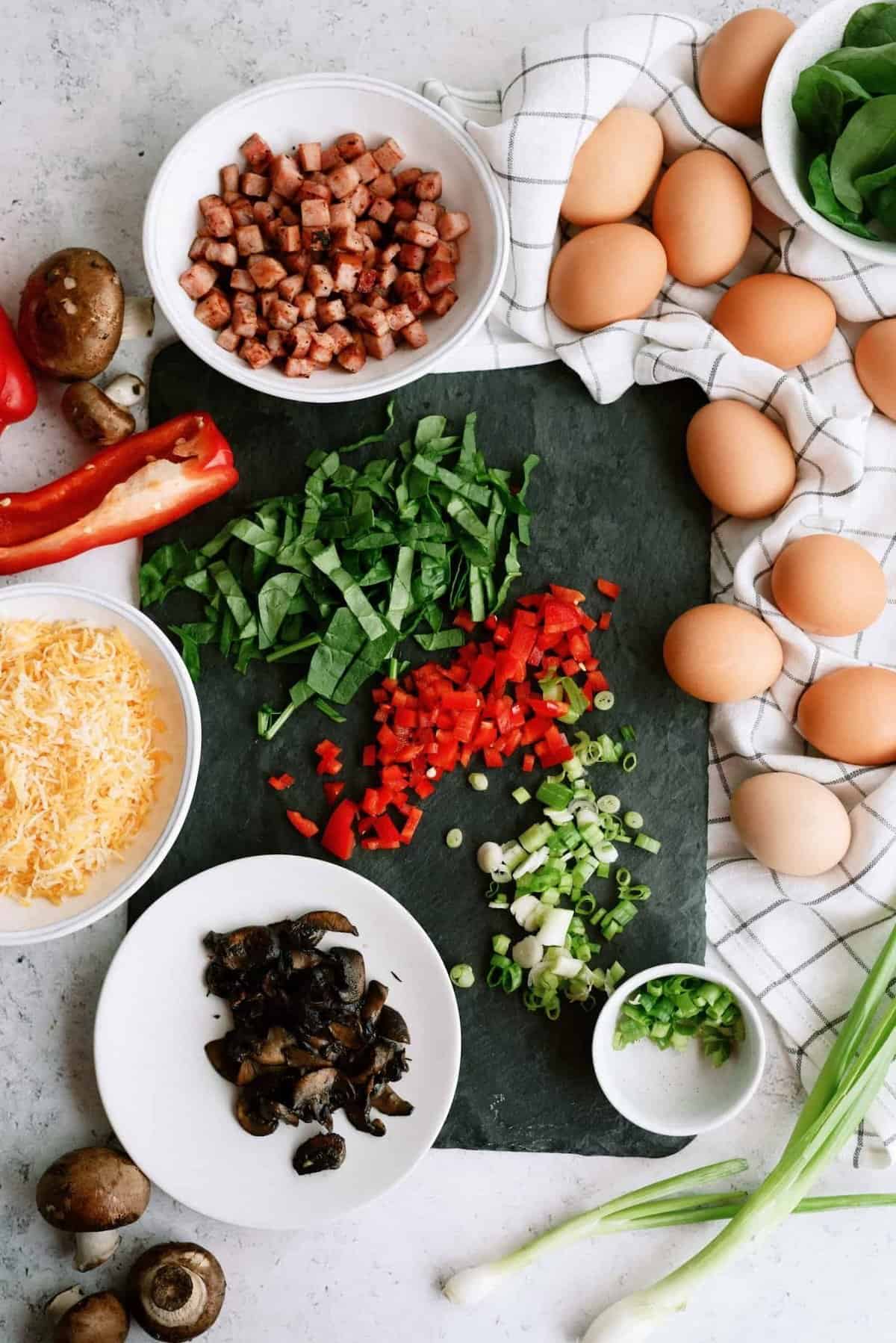 Chopped vegetables, diced bacon, grated cheese, and whole eggs are arranged on a table with a black slate in the center, surrounded by mushrooms, spinach, and a red bell pepper.