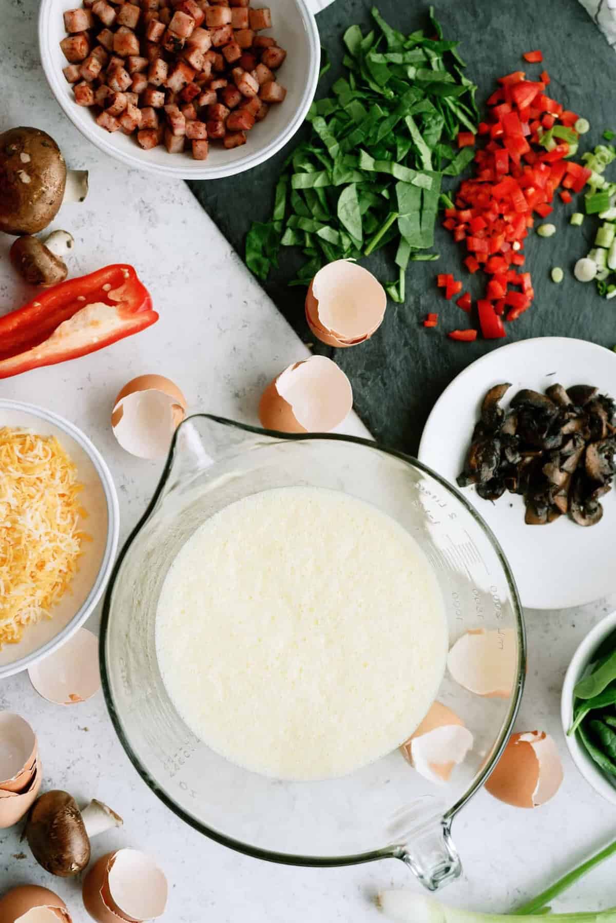 Various toppings including diced bacon, chopped spinach, red peppers, mushrooms, and shredded cheese surround a bowl of beaten eggs, with scattered eggshells nearby.