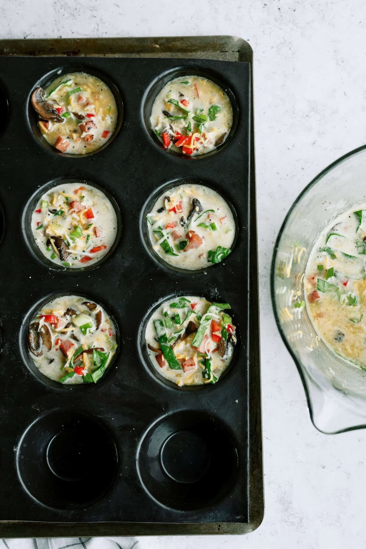 Muffin tray filled with uncooked egg mixture containing vegetables like bell peppers, mushrooms, and greens beside an empty section and a bowl of remaining mixture on a white counter.