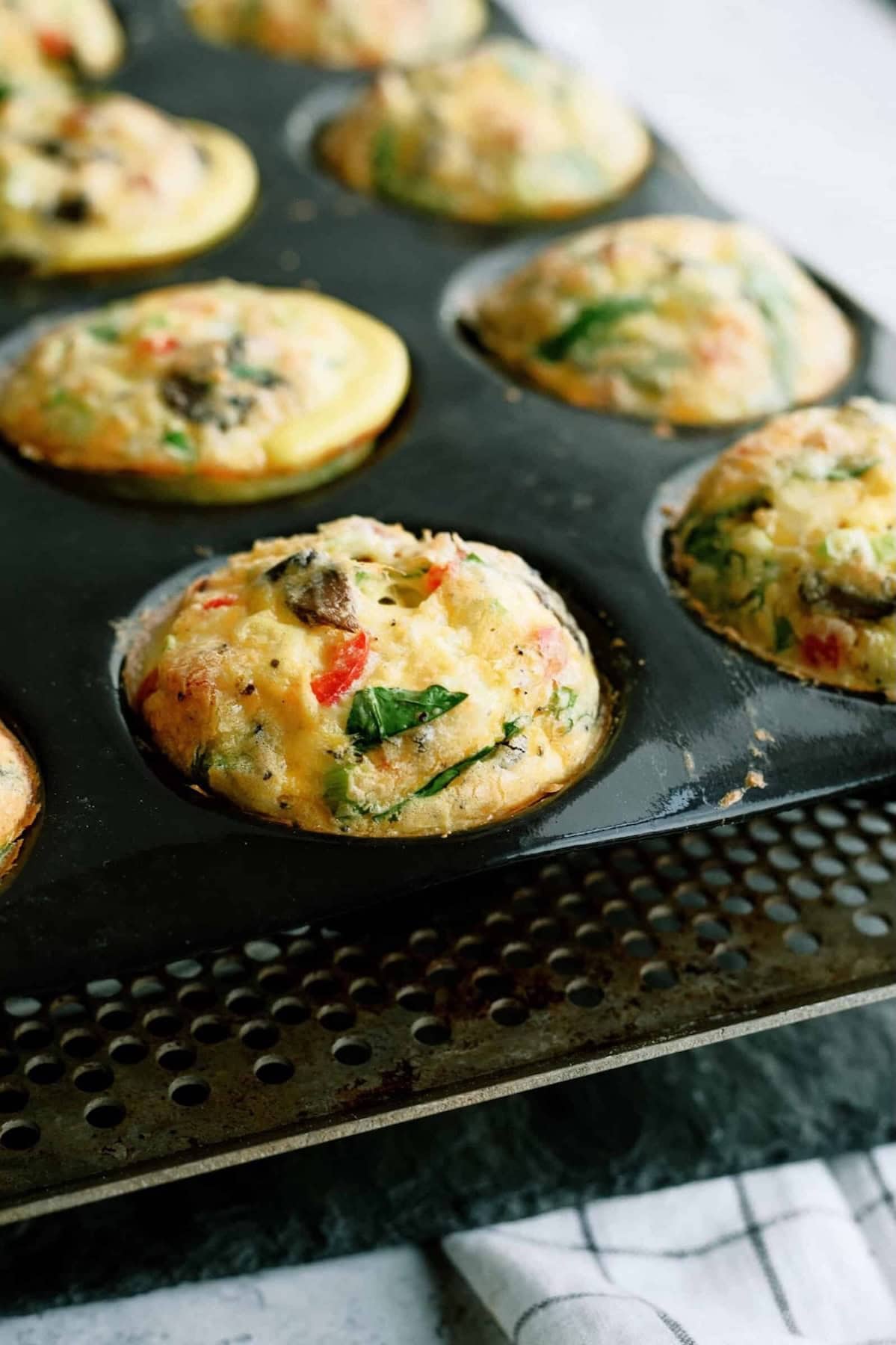 Muffin tin filled with vegetable egg muffins, featuring visible spinach, red peppers, and mushrooms.