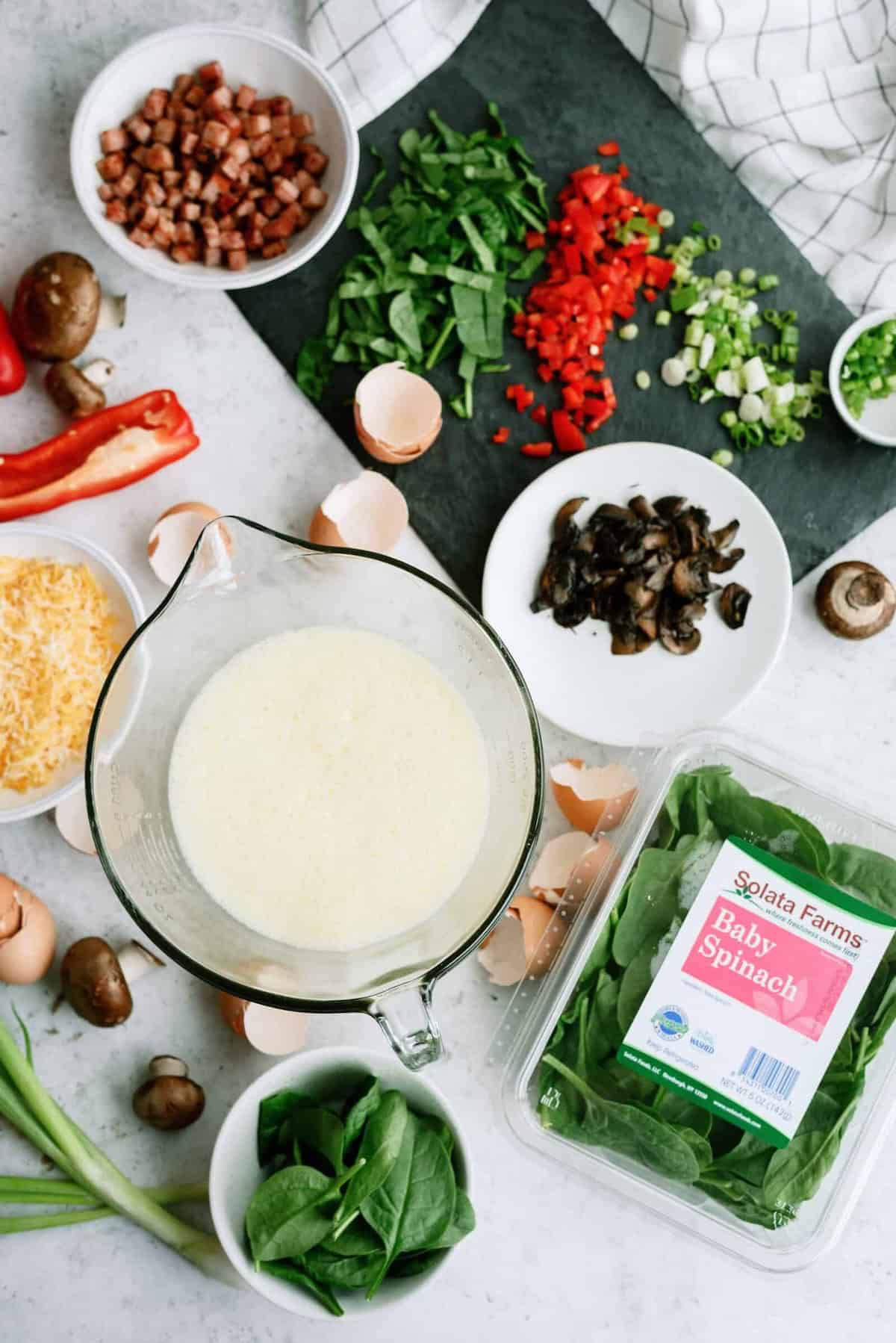 A kitchen counter with chopped vegetables, a measuring cup of liquid, a carton of baby spinach, cheese, and empty eggshells. Ingredients likely for a cooking preparation.