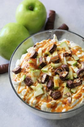 A bowl of caramel apple salad garnished with chopped pieces of apples and chocolate bars. Two green apples and pieces of chocolate bars are placed next to the bowl.