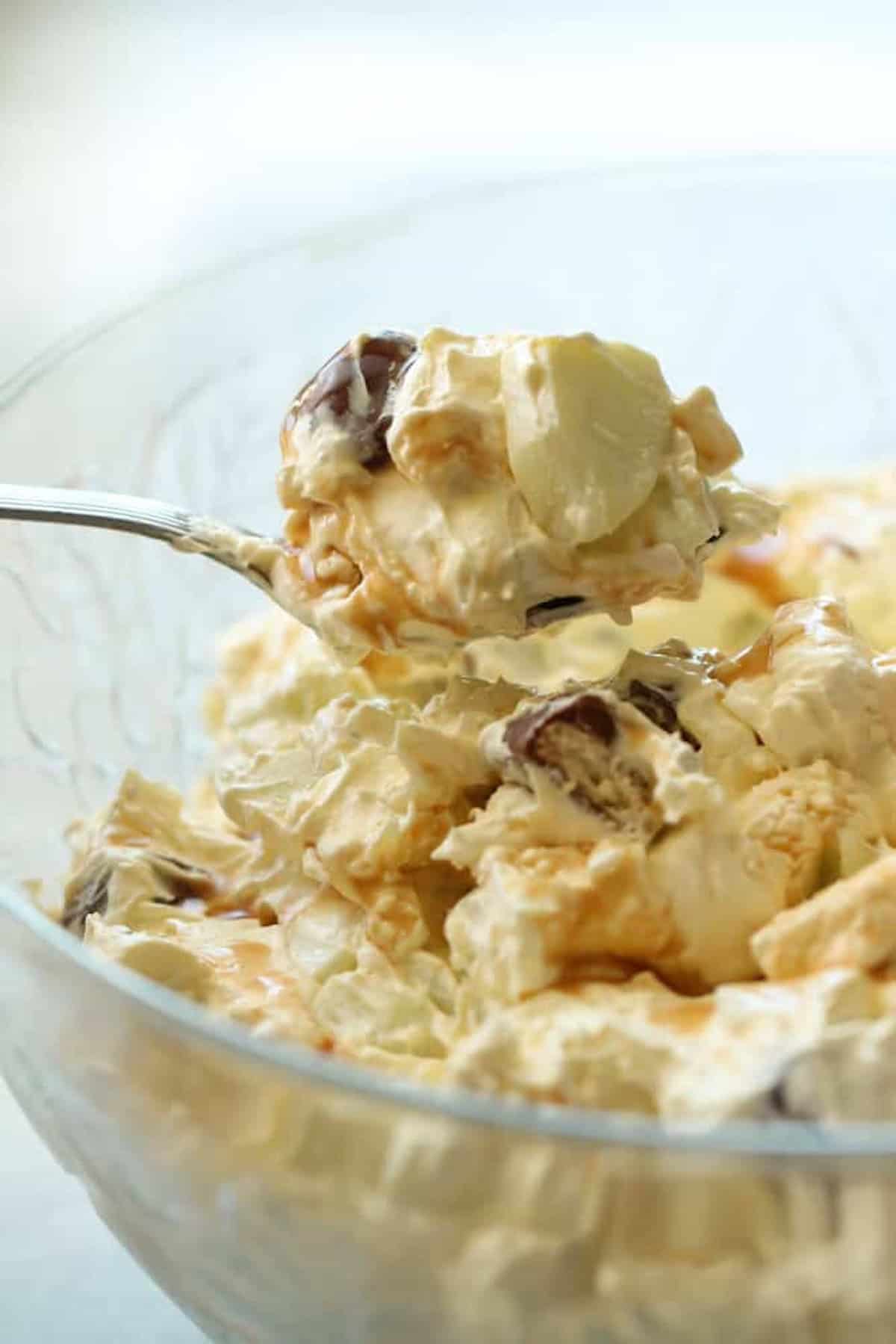 A spoonful of creamy dessert with whipped topping, grapes, and fruit pieces is held above a glass bowl filled with the same mixture.