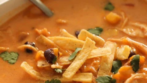 A bowl of creamy tortilla soup with tortilla strips, black beans, corn, cilantro, and a spoon.