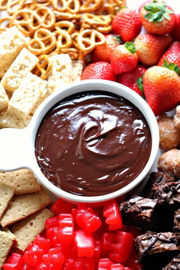 Assorted snacks including strawberries, brownies, pretzels, Rice Krispies treats, and gummy candies surround a bowl of chocolate dip.