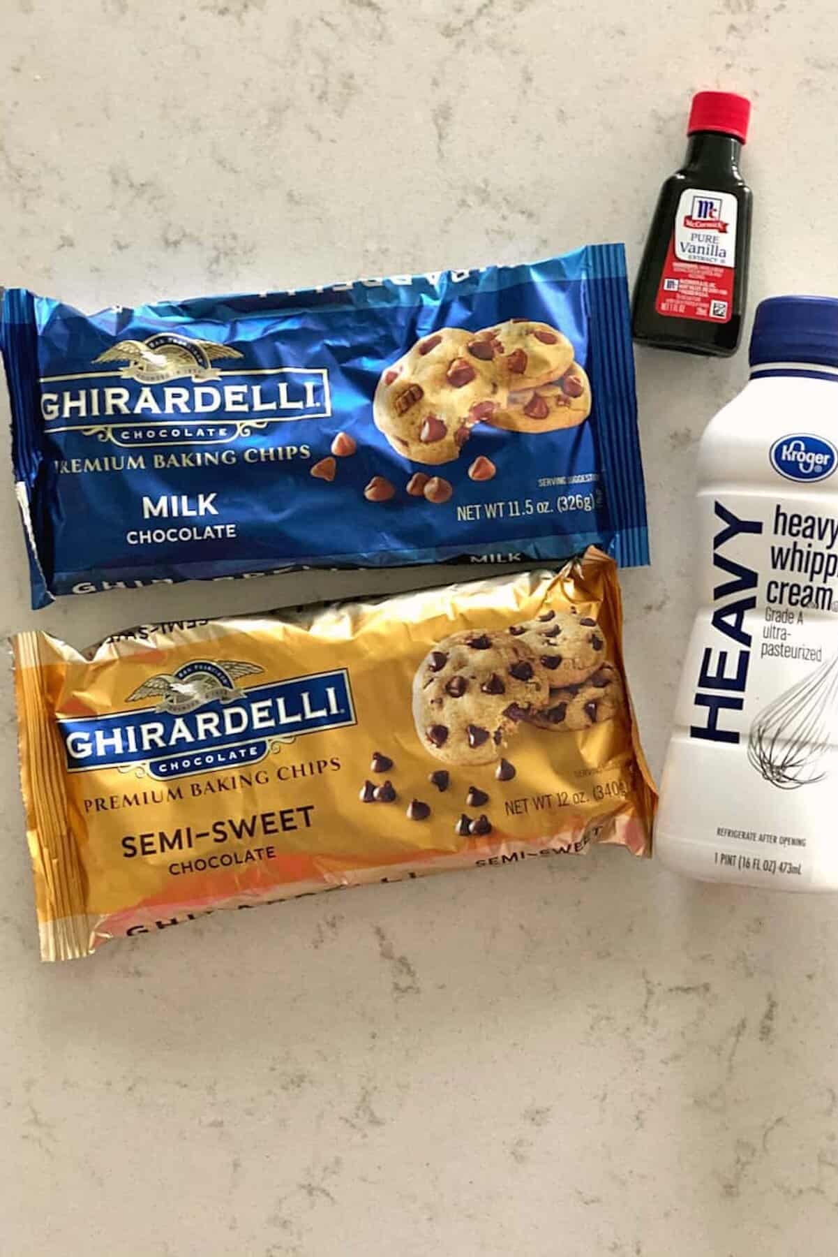Two bags of Ghirardelli chocolate chips, a bottle of heavy whipping cream, and a small bottle of vanilla extract are arranged on a light surface.