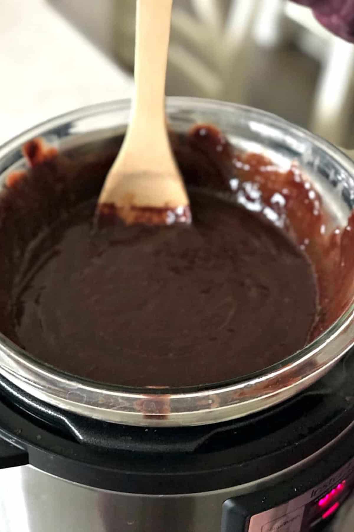 A wooden spoon stirs melted chocolate in a glass bowl set over an electric pressure cooker.