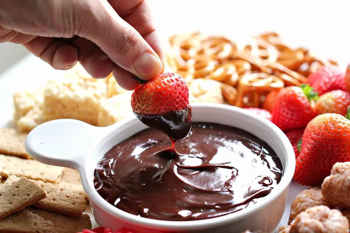 A hand dips a strawberry into a bowl of melted chocolate surrounded by pretzels, graham crackers, and other snacks.