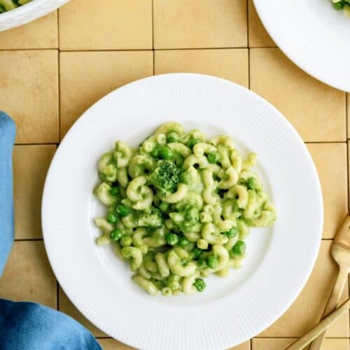 A white plate of pasta with green sauce and vegetables, placed on a yellow tiled surface next to gold cutlery and a blue cloth.