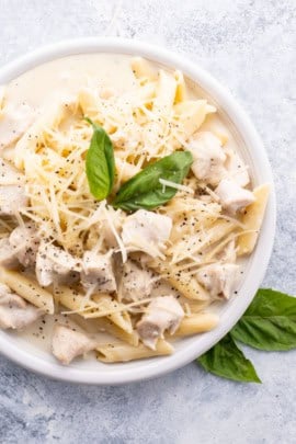 A white plate filled with creamy pasta and pieces of chicken, topped with grated cheese, cracked black pepper, and garnished with a fresh basil leaf.