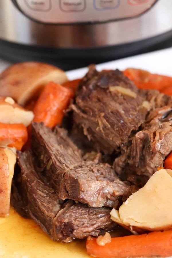 A plate of pot roast with chunks of beef, carrots, and potatoes in front of an electric pressure cooker.