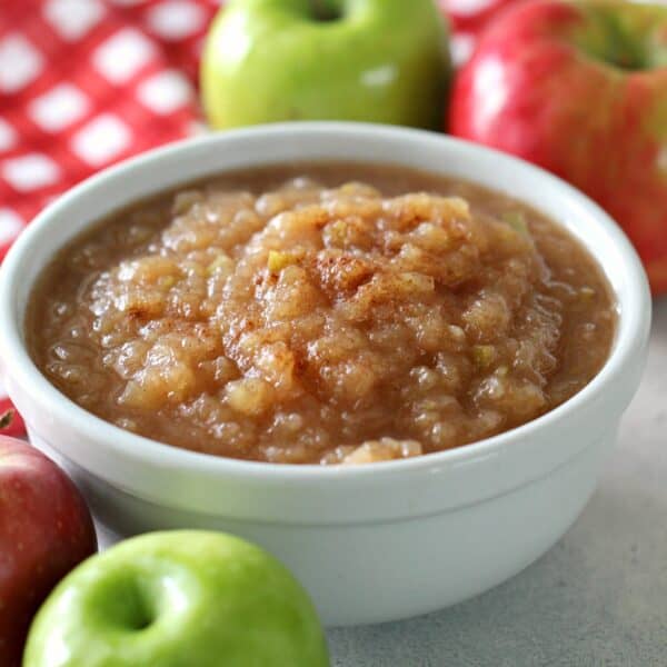 A bowl of applesauce is surrounded by whole red and green apples, with a red and white checkered cloth in the background.