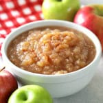 A bowl of applesauce is surrounded by whole red and green apples, with a red and white checkered cloth in the background.