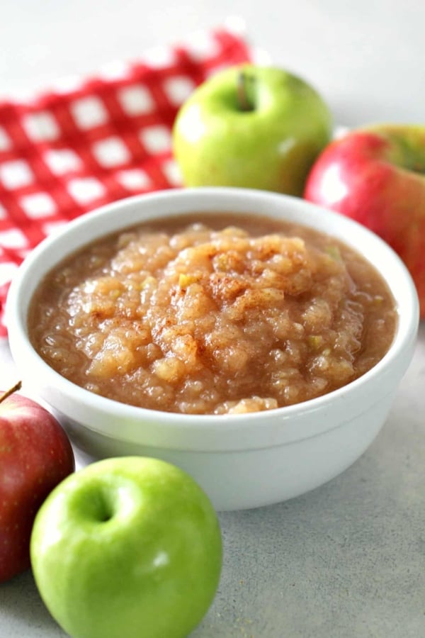 A bowl of applesauce is surrounded by whole red and green apples, with a red and white checkered cloth in the background.