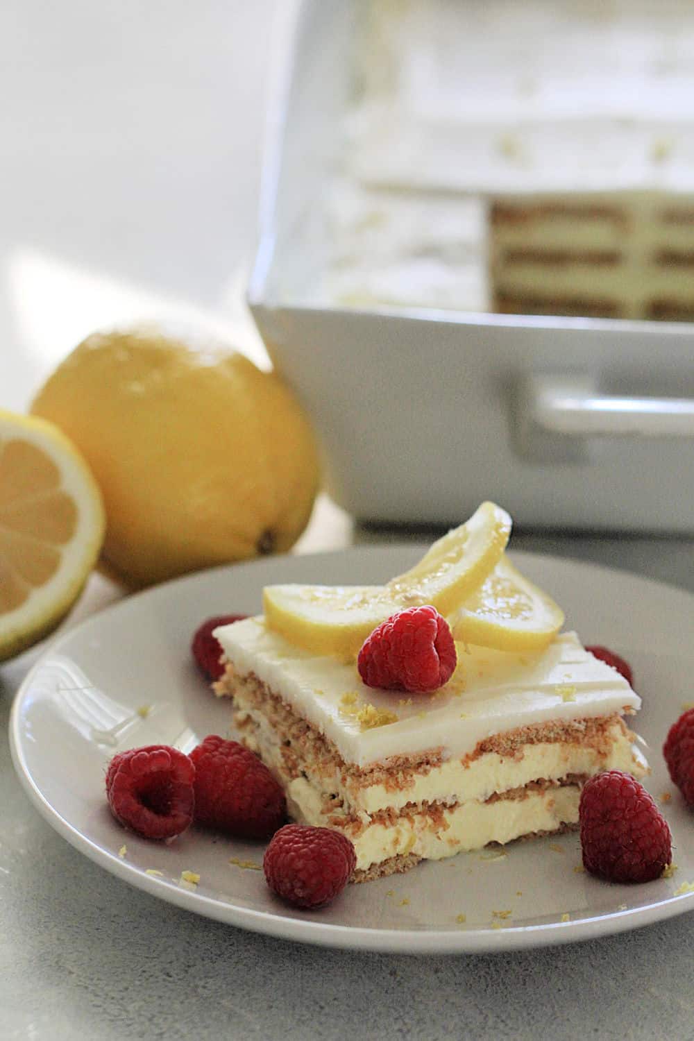 No Bake Lemon Ice Box Cake Recipe (Eclair Cake) on a white plate topped with lemon slices and raspberries
