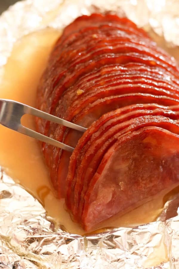 A metal fork holds a sliced ham that is arranged on a piece of foil, covered in a glossy glaze, with juices pooling around it.