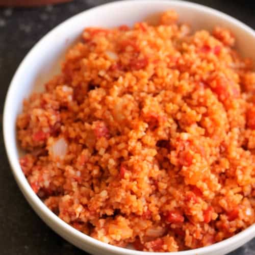 A white bowl filled with cooked Spanish cauliflower rice mixed with tomatoes and onions sits on a dark countertop.