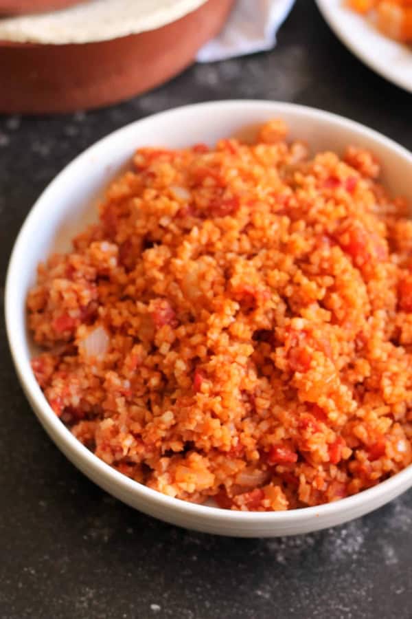A white bowl filled with cooked Spanish cauliflower rice mixed with tomatoes and onions sits on a dark countertop.