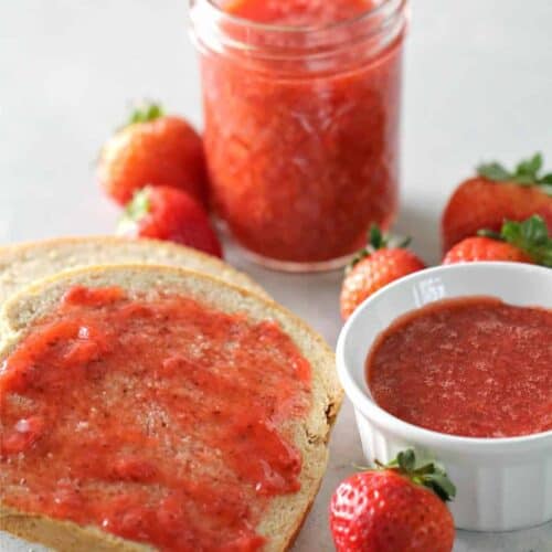 A slice of bread with strawberry jam, a small bowl and a jar of jam, and several fresh strawberries on a light surface.