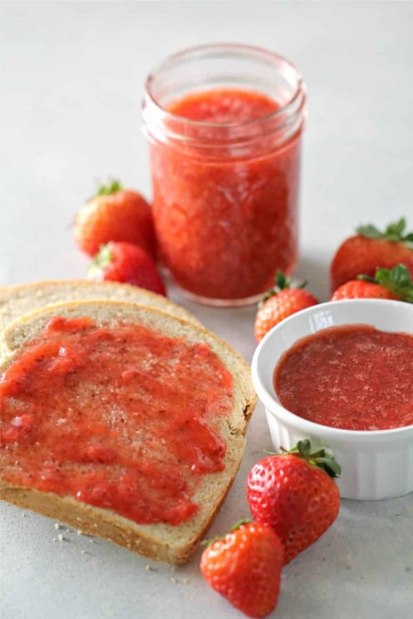 A slice of bread with strawberry jam, a small bowl and a jar of jam, and several fresh strawberries on a light surface.