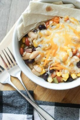A bowl of mixed beans, corn, and melted cheese with folded tortillas on the side. Two forks are placed on a wooden surface next to a checkered napkin.