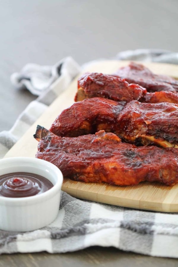 Several pieces of barbecued ribs on a wooden cutting board with a small white dish of barbecue sauce next to them, placed on a checkered cloth.