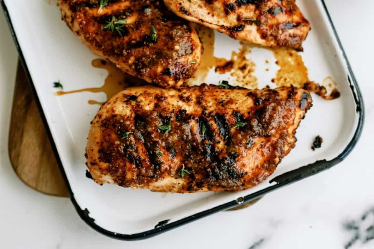 Grilled chicken breasts with visible grill marks and seasoning, garnished with herbs, served on a white enamel tray.