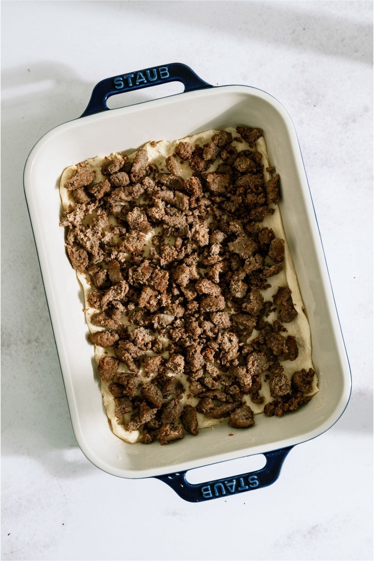 Browned and crumbled sausage on top of biscuit dough in casserole dish.