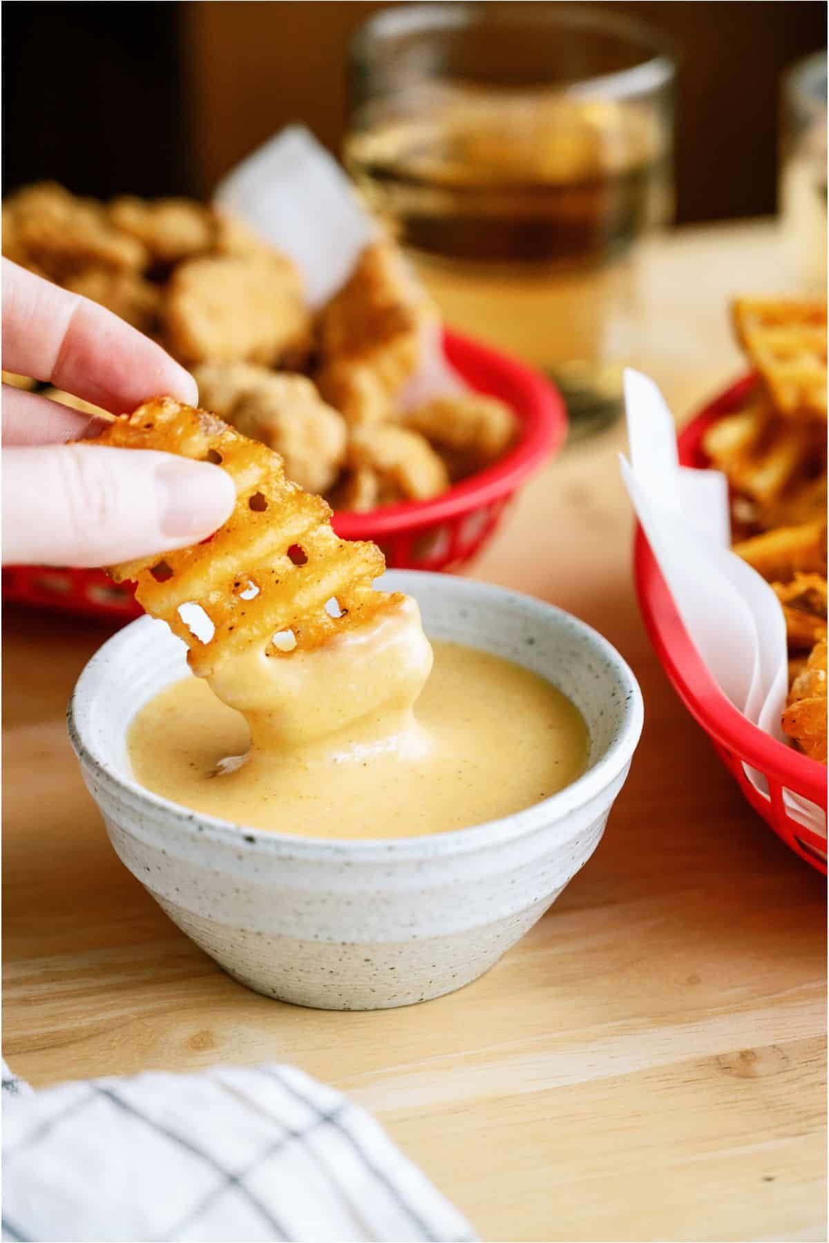 A hand dips a waffle fry into a bowl of creamy sauce, with baskets of fried food visible in the background on a wooden table.