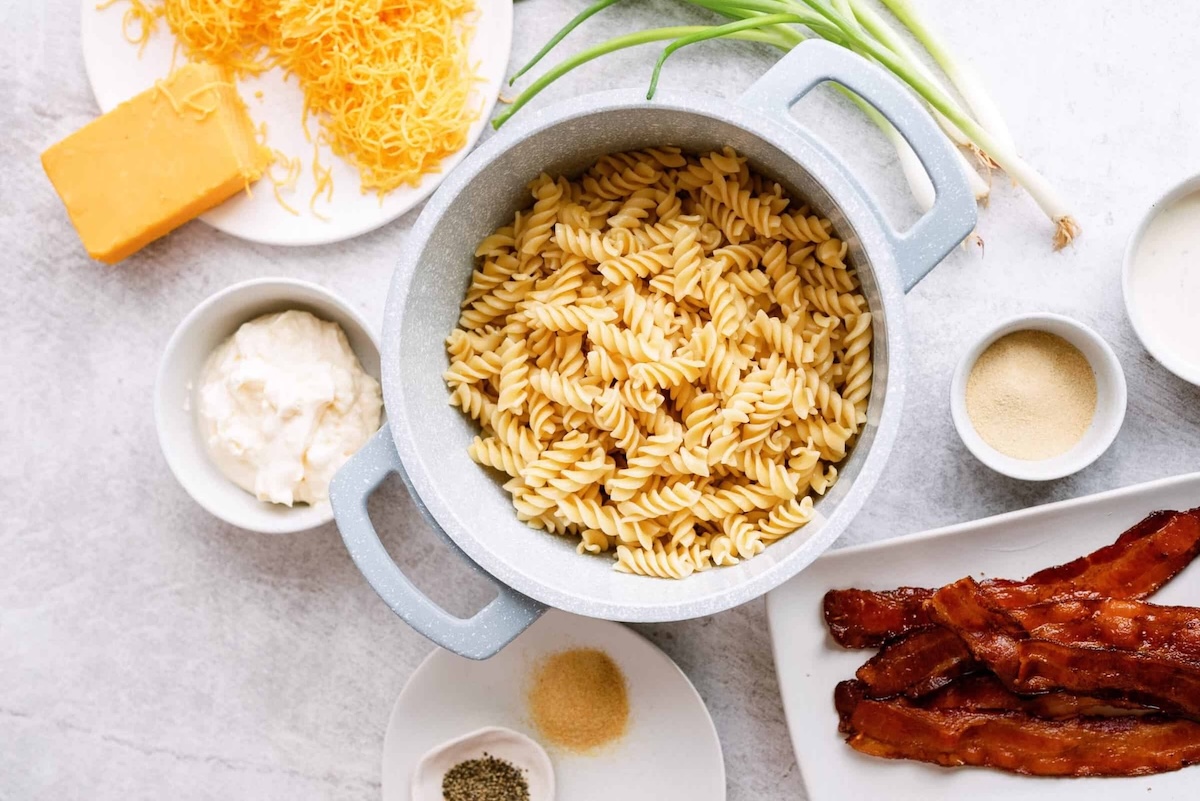A pot of cooked rotini pasta surrounded by cheese, green onions, mayonnaise, spices, and cooked bacon on a light surface.