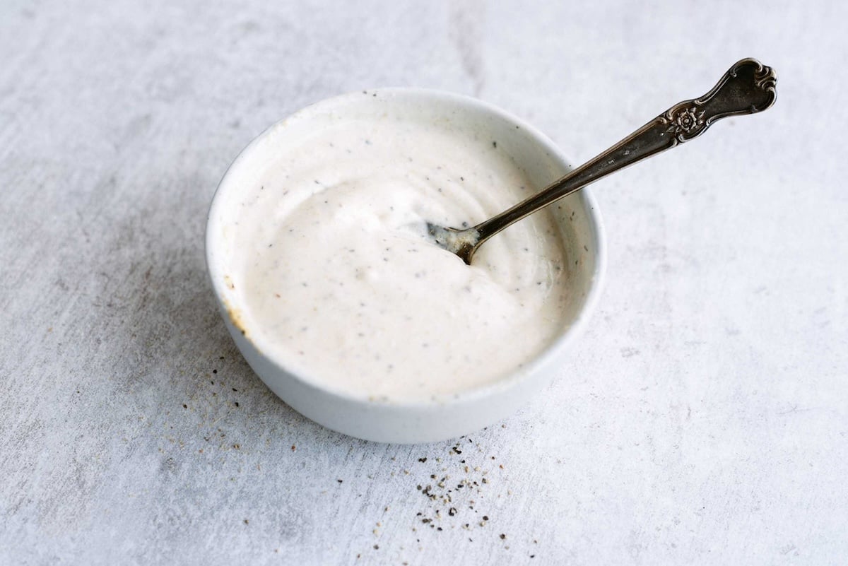 A small white bowl contains creamy white sauce with visible seasoning, and a spoon resting inside. The bowl is placed on a light gray surface.