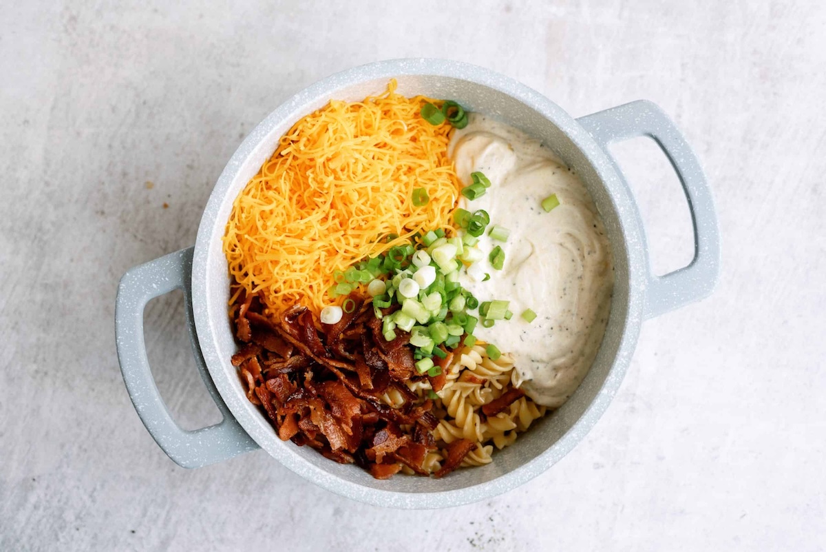 A pot containing shredded cheese, chopped bacon, pasta, creamy sauce, and sliced green onions on a light surface.